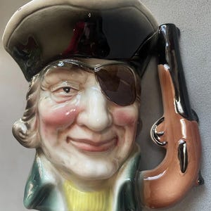 May include: A ceramic pirate-themed mug with a smiling face, eye patch, and tricorn hat. The mug is detailed with a brown pistol handle. The pirate's face has rosy cheeks and a yellow sweater.