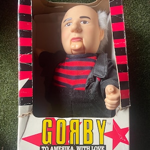 May include: A vintage Goaby doll in its original box. The doll has a bald head, white hair, and is wearing a black jacket over a red and black striped shirt. The box has red and black stripes and the text "GOABY TO AMERIKA, WITH LOVE."