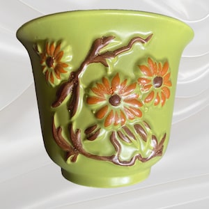 May include: A lime green ceramic planter with embossed floral and stem designs in orange and brown. The planter has a flared rim and a rounded base, suitable for small plants or succulents.