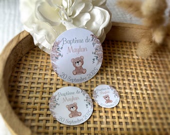 May include: Three round stickers with the text "Baptême de Maylan" and "20 Septembre 2025" in a decorative floral design, featuring a teddy bear illustration. The stickers are in a woven tray with a white flower and dried plants.
