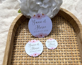 May include: Three round stickers with the text "Yann et Florie 25 JUILLET 2026" in elegant script, surrounded by pink floral accents. A large white flower and a woven tray are in the background.