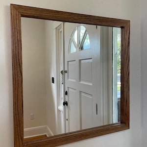 以下が含まれることがあります： 茶色の木製フレームの長方形の鏡。鏡は、アーチ型の窓と屋外の様子が映る白いドアを映しています。フレームには木目がはっきりと見えます。