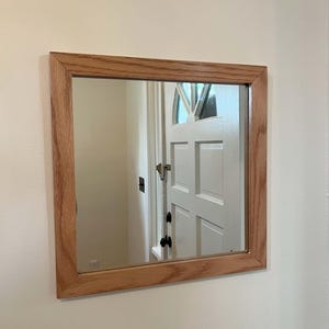 May include: A square mirror with a light wood frame. The mirror is reflecting a white door with a black door handle.