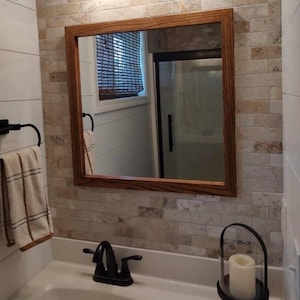 Puede incluir: Un espejo de pared cuadrado con un marco de madera de tono medio montado en una pared con azulejos de piedra rectangulares claros. El espejo refleja una ventana con una persiana de bambú marrón y una puerta de ducha con un tirador negro.