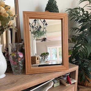 May include: A rectangular mirror with a light brown wood grain frame. The mirror reflects a room interior featuring a black chandelier, a ceiling fan, and a window. The frame has a beveled edge design and a smooth, natural wood finish.