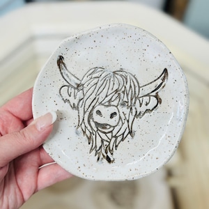 May include: A white ceramic dish with a speckled glaze features a black and white illustration of a Highland cow's face.