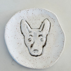 May include: White ceramic dish with a speckled glaze. The dish has a black line drawing of a dog's face on it.