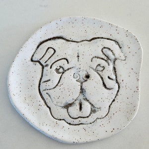 May include: A white ceramic dish with a speckled surface, featuring a detailed outline of a bulldog's face. The dog's features are etched in dark brown, creating a charming, rustic aesthetic. This decorative dish is ideal for pet lovers.