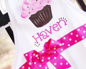 Peut inclure: Tablier blanc avec un motif de cupcake rose et marron, un nœud rose à pois blancs et le nom "Haven" écrit en rose.