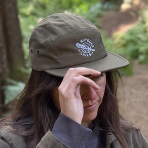 May include: An olive green five-panel cap featuring a white embroidered graphic on the front. The design depicts a hand holding a pipe wrench with the text "HAYDUKE LIVES" arched around the illustration. The cap has a flat brim and side eyelets.