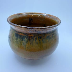 Pode incluir: Uma tigela de cerâmica artesanal, com formato largo e arredondado. A tigela apresenta um acabamento brilhante com uma mistura de tons castanhos, dourados e verdes. O interior é de um tom mais escuro, criando um contraste. Um objeto decorativo para a casa, com cerca de 15 cm de diâmetro.