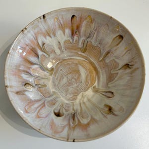 Puede incluir: Un cuenco de cerámica hecho a mano con un esmalte único. El cuenco presenta una base de color crema con un diseño en espiral en el centro. El borde y la superficie exterior tienen un patrón fluido de melocotón, marrón y blanco. El cuenco es una pieza decorativa.