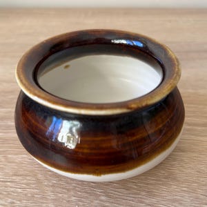 Può includere: Una piccola ciotola rotonda in ceramica con esterno marrone lucido e interno bianco. Il bordo è marrone chiaro e la ciotola poggia su una base bianca. La ciotola è probabilmente fatta a mano e adatta per servire salse o piccole porzioni.