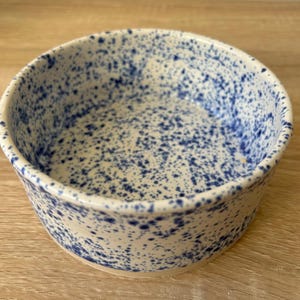 Puede incluir: Un cuenco redondo de cerámica blanquecina con un patrón azul moteado. El cuenco tiene un diseño sencillo y clásico, adecuado para servir alimentos o como pieza decorativa. Las motas azules están distribuidas uniformemente.