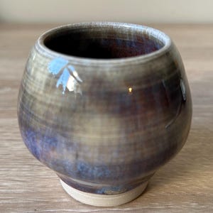 May include: A handcrafted ceramic cup with a rounded form and a small, flat base. The cup displays a mix of brown, blue, and gold hues, with a glossy surface. The inside is a deep reddish-brown. A unique piece for drinks.