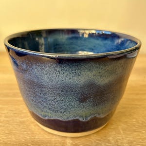 Puede incluir: Un cuenco de cerámica hecho a mano con un degradado de tonos azules. El cuenco pasa de un índigo profundo en la base a un azul más claro y moteado hacia el centro, y un borde azul oscuro. El interior es una superficie brillante y reflectante.