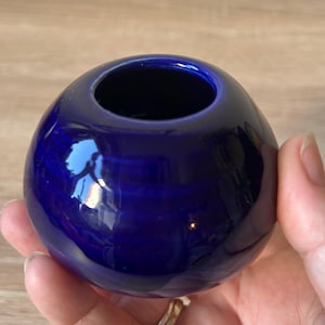 May include: A small, round, cobalt blue ceramic vase with a glossy finish. The vase has a single opening at the top and reflects light. The vase is held in a hand.