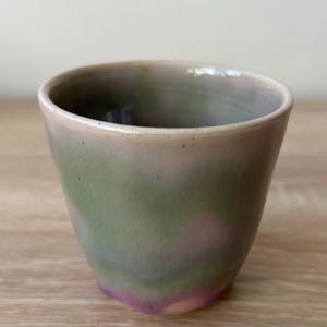 Könnte beinhalten: Eine handgefertigte Keramiktasse mit einem einzigartigen Farbverlauf. Die Tasse wechselt von einem zarten Rosa am Rand zu einer grünen Mitte und endet mit einem Hauch von Lila am Boden. Die Tasse hat eine glatte, leicht strukturierte Oberfläche.