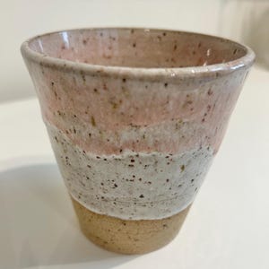 Puede incluir: Una taza de cerámica con un esmalte moteado en tonos de rosa, blanco y marrón. La taza tiene una forma redondeada y un borde ancho.