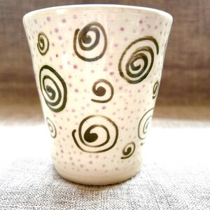 Può includere: Un bicchiere in ceramica con base color crema, decorato con disegni a spirale verde scuro e piccoli pois viola. La tazza è cilindrica e poggia su una superficie di colore neutro.