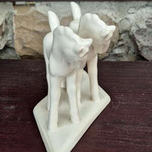 Pode incluir: Uma estatueta de cer&acirc;mica creme retratando tr&ecirc;s veados estilizados. Os veados est&atilde;o em p&eacute; sobre uma base triangular. A estatueta tem um acabamento liso e brilhante e est&aacute; colocada sobre uma superf&iacute;cie de madeira escura e uma parede de pedra.