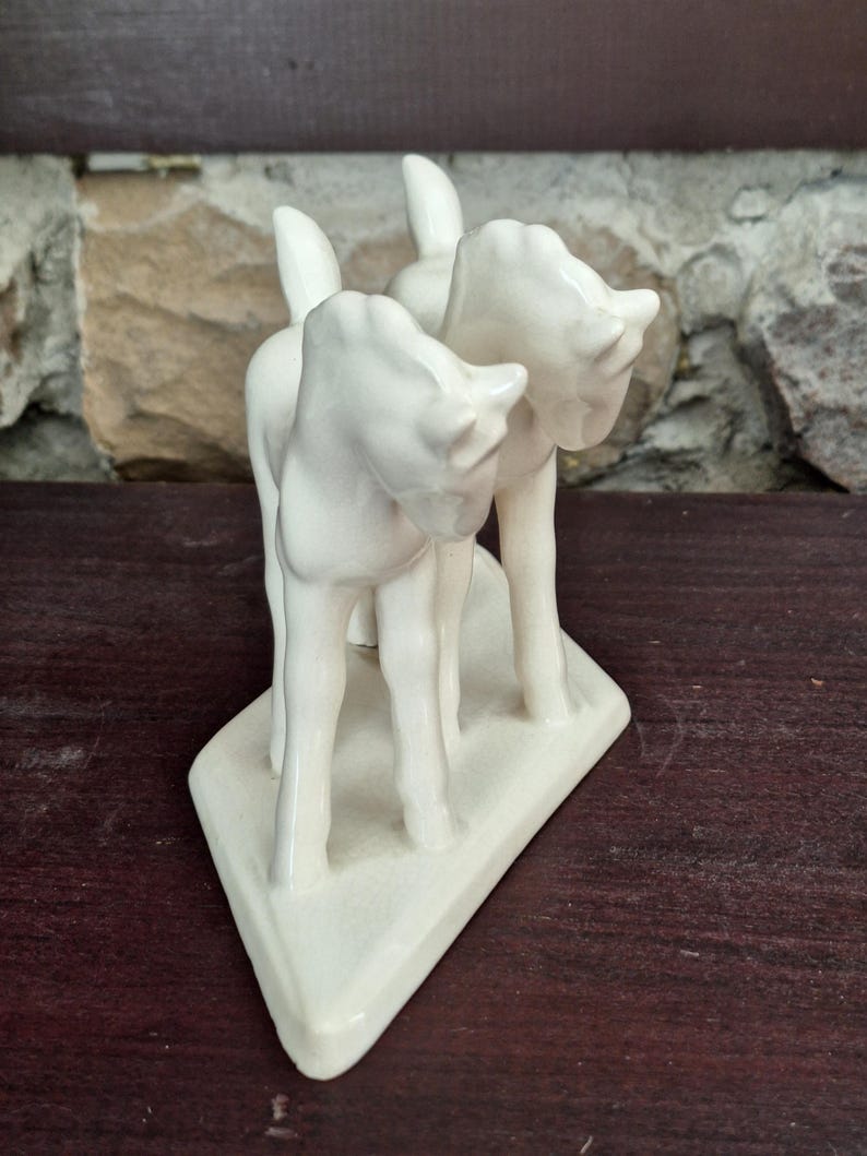 Pode incluir: Uma estatueta de cer&acirc;mica creme retratando tr&ecirc;s veados estilizados. Os veados est&atilde;o em p&eacute; sobre uma base triangular. A estatueta tem um acabamento liso e brilhante e est&aacute; colocada sobre uma superf&iacute;cie de madeira escura e uma parede de pedra.