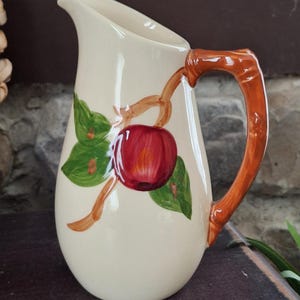 May include: Cream-colored ceramic pitcher with a red apple and green leaf design. The handle is a bamboo-style design in a warm brown color. The pitcher is a classic shape, perfect for serving beverages.