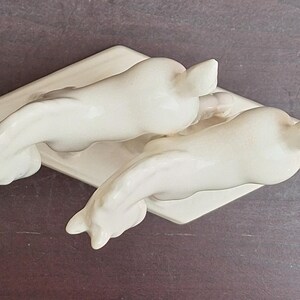 Pode incluir: Duas estatuetas de golfinhos de cer&acirc;mica esbranqui&ccedil;ada, cada uma com um corpo curvo e cauda para cima, repousando sobre uma base retangular. Os golfinhos est&atilde;o posicionados em dire&ccedil;&otilde;es opostas, criando um design sim&eacute;trico.