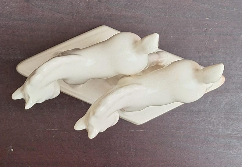 Pode incluir: Duas estatuetas de golfinhos de cer&acirc;mica esbranqui&ccedil;ada, cada uma com um corpo curvo e cauda para cima, repousando sobre uma base retangular. Os golfinhos est&atilde;o posicionados em dire&ccedil;&otilde;es opostas, criando um design sim&eacute;trico.