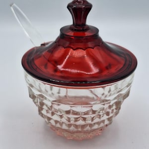 May include: A clear glass sugar bowl with a ruby red lid and handle. The bowl has a diamond pattern, and a clear spoon rests on the side. The lid has a decorative knob.