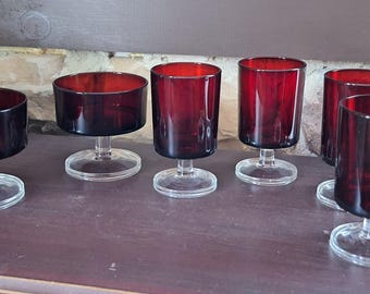 May include: A set of six vintage ruby red glass goblets. The glasses have a cylindrical bowl and a clear, circular base. The goblets are arranged on a dark brown surface, with a stone wall in the background.