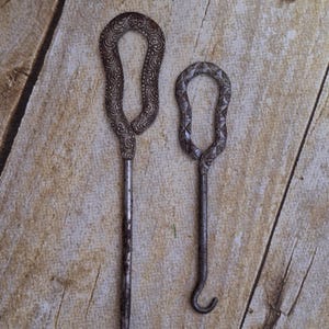 May include: Two antique metal shoe horns with ornate designs. The larger horn has a floral pattern, while the smaller horn has a geometric pattern. Both horns have a hook at the end.