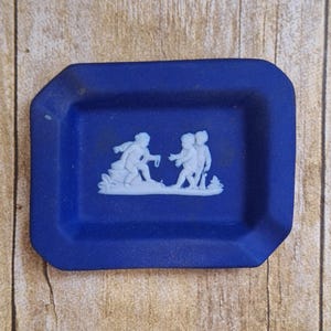 May include: A cobalt blue rectangular trinket dish with a white bas-relief design of classical figures. The dish has a slightly textured surface and is set against a wooden background.