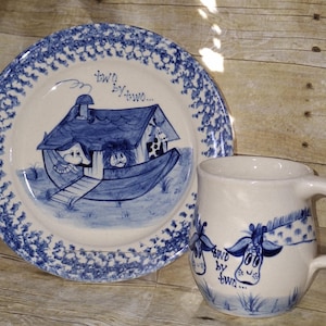 May include: A white ceramic mug and plate set with blue illustrations. The plate features a blue drawing of Noah's Ark with animals, and the text "two by two." The mug has a giraffe design and the same text.