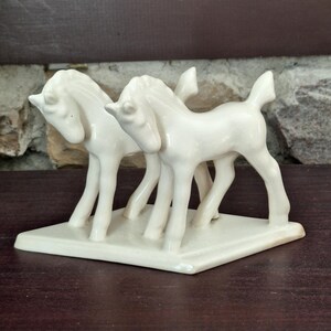 Pode incluir: Uma estatueta de cer&acirc;mica creme com dois cavalos estilizados em p&eacute; lado a lado sobre uma base retangular. Os cavalos t&ecirc;m pernas finas, pesco&ccedil;os curvos e crinas detalhadas. O objeto &eacute; decorativo.