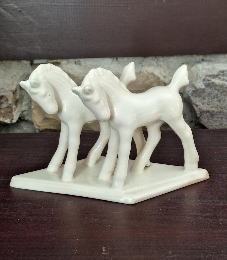 Pode incluir: Uma estatueta de cer&acirc;mica creme com dois cavalos estilizados em p&eacute; lado a lado sobre uma base retangular. Os cavalos t&ecirc;m pernas finas, pesco&ccedil;os curvos e crinas detalhadas. O objeto &eacute; decorativo.