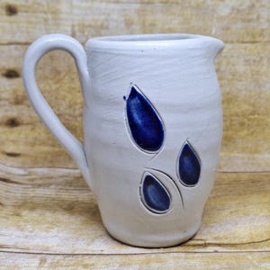 May include: White ceramic pitcher with a curved handle and spout. The pitcher features three blue teardrop-shaped designs on the front. The handmade pottery has a textured surface and is set against a wooden background.