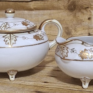 May include: A white porcelain sugar bowl and creamer set with gold leaf accents. The set features a delicate floral design with leaves and a gold trim.