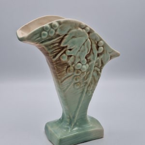 May include: A light green ceramic vase with a flared, asymmetrical rim and a rectangular base. The exterior features a raised, molded relief pattern of grape clusters and leaves, with brown shading highlighting the textured details of the design.