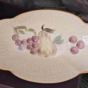 May include: Cream-colored ceramic serving platter with a scalloped edge and gold trim. The platter features a painted design of a pear, grapes, and cherries. The words "Breads", "Biscuits", "Rolls", and "Buns" are embossed on the platter.