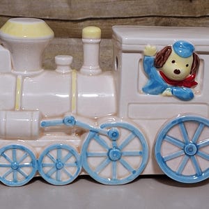 May include: A white ceramic toy train with a yellow chimney and blue wheels. A cartoon dog wearing a blue hat and red scarf is in the caboose window.