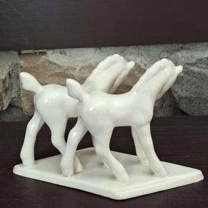 Pode incluir: Estatueta de cer&acirc;mica creme com dois cavalos estilizados. Os cavalos est&atilde;o em p&eacute; sobre uma base retangular, com a cabe&ccedil;a levantada. A estatueta tem um acabamento brilhante e &eacute; provavelmente um item decorativo.