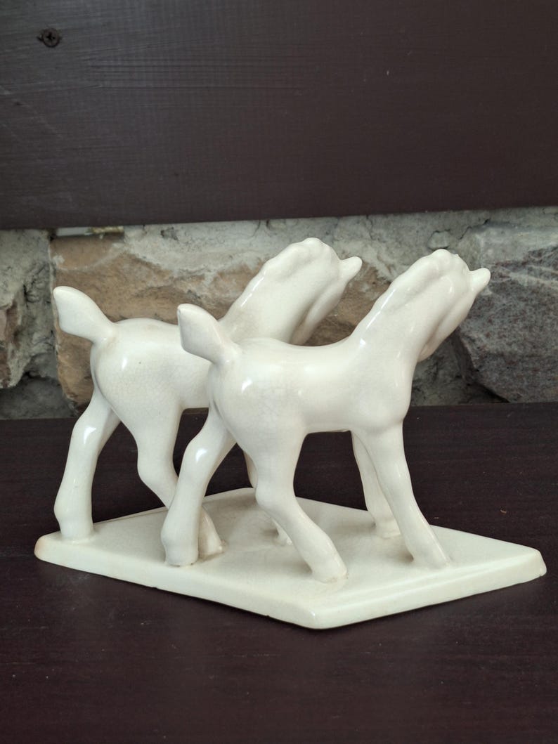 Pode incluir: Estatueta de cer&acirc;mica creme com dois cavalos estilizados. Os cavalos est&atilde;o em p&eacute; sobre uma base retangular, com a cabe&ccedil;a levantada. A estatueta tem um acabamento brilhante e &eacute; provavelmente um item decorativo.
