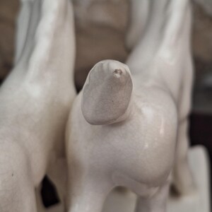 Pode incluir: Um close-up de uma estatueta de cavalo de cer&acirc;mica branca. A estatueta retrata dois cavalos, com a parte traseira de um cavalo em foco n&iacute;tido. A cer&acirc;mica tem uma apar&ecirc;ncia ligeiramente envelhecida.