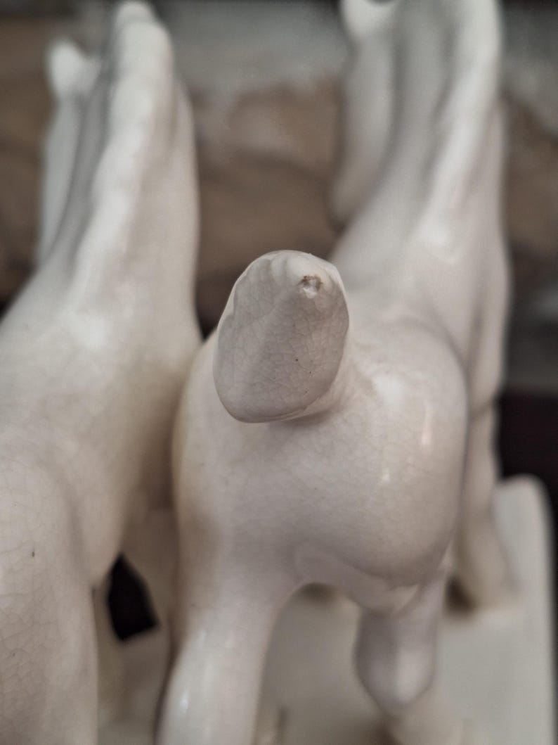 Pode incluir: Um close-up de uma estatueta de cavalo de cer&acirc;mica branca. A estatueta retrata dois cavalos, com a parte traseira de um cavalo em foco n&iacute;tido. A cer&acirc;mica tem uma apar&ecirc;ncia ligeiramente envelhecida.