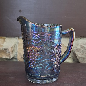 May include: An iridescent blue glass pitcher with a grape design. The pitcher has a handle and a scalloped rim. The glass reflects a rainbow of colors, including purple, gold, and green. The pitcher is likely vintage and suitable for serving beverages.