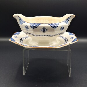 May include: A white gravy boat and matching saucer set. The boat has a curved handle and a flared rim. Both pieces are decorated with blue floral and geometric patterns and a gold trim. The set is displayed on a clear acrylic stand.