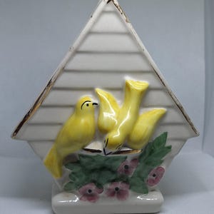 May include: A ceramic birdhouse-shaped figurine with a white background and a triangular roof. Two yellow birds are perched on a branch with green leaves and pink flowers. The roof has horizontal lines and gold trim.