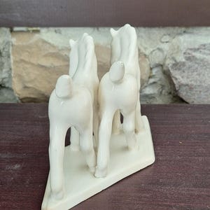 Pode incluir: Estatueta de cavalo de cer&acirc;mica creme. Quatro cavalos em p&eacute; juntos em uma base retangular. Os cavalos s&atilde;o vistos por tr&aacute;s. A estatueta tem um acabamento brilhante. A base &eacute; retangular.