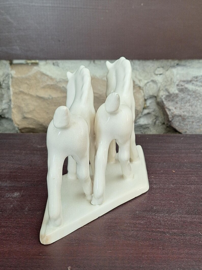 Pode incluir: Estatueta de cavalo de cer&acirc;mica creme. Quatro cavalos em p&eacute; juntos em uma base retangular. Os cavalos s&atilde;o vistos por tr&aacute;s. A estatueta tem um acabamento brilhante. A base &eacute; retangular.